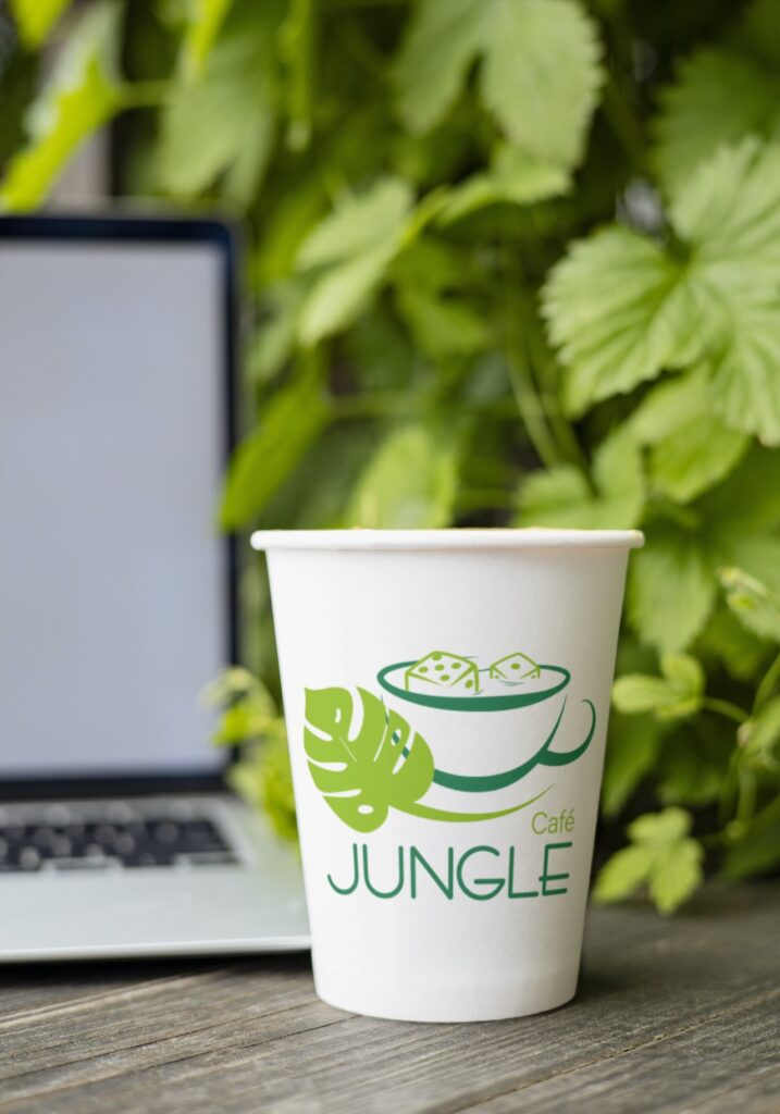 Verre en carton avec un logo de café sur la jungle posé sur une table en bois à côté d'un ordinateur et devant un mur végétal